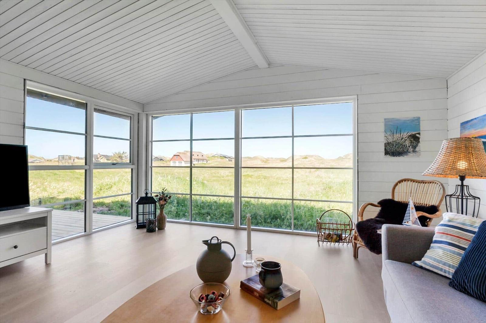 Wohnzimmer mit großen Fenstern, Blick auf Grasland und Dächer. Sofa, Couchtisch und Lampe sichtbar.