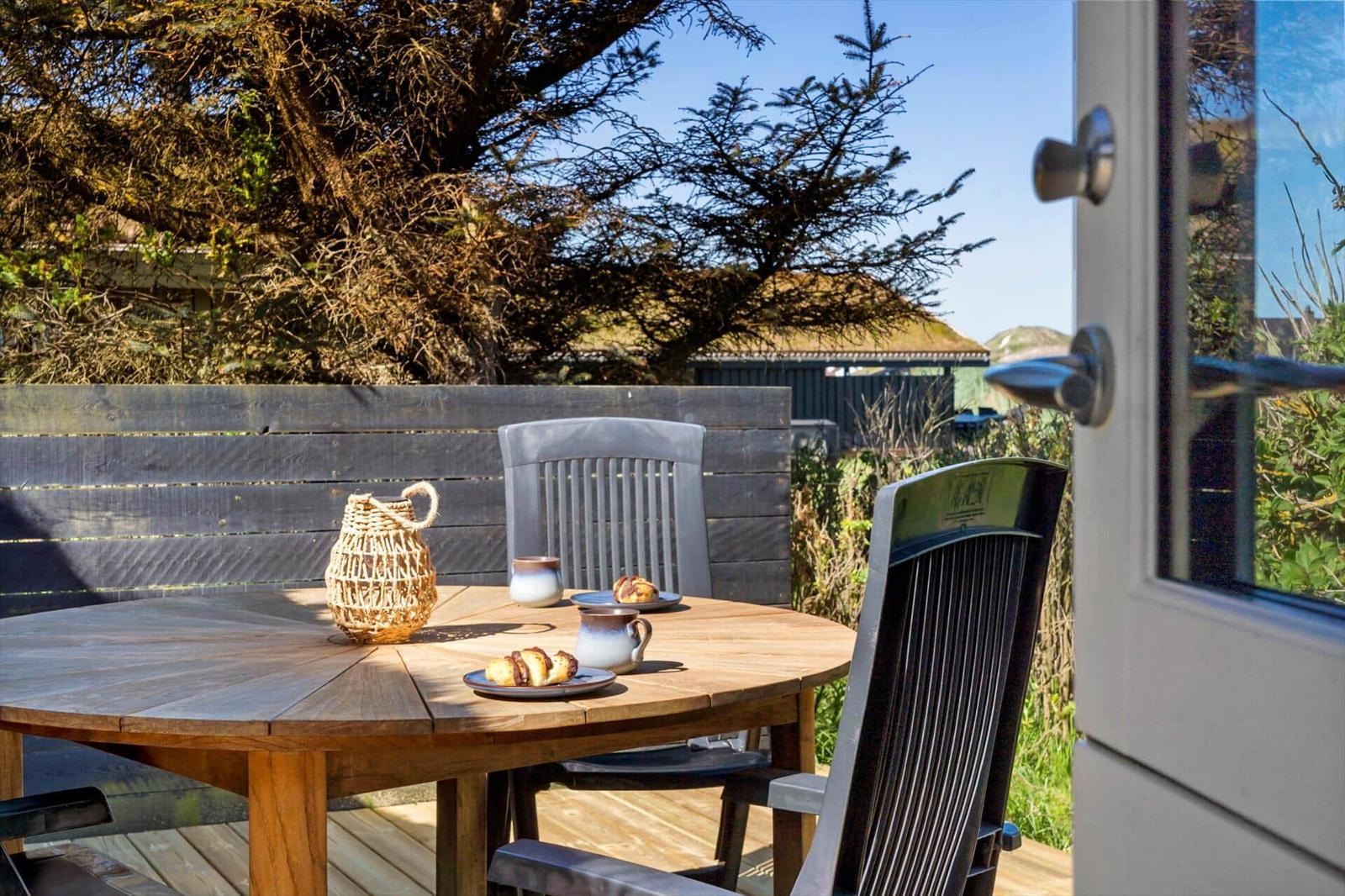 Holztisch mit Stühlen und Frühstück auf Terrasse unter Bäumen.