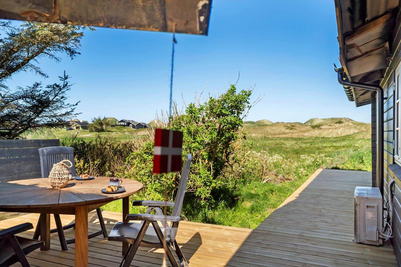 Terrasse mit Tisch, Stühlen und dänischer Flagge. Blick auf grüne Wiesen und Hügel.