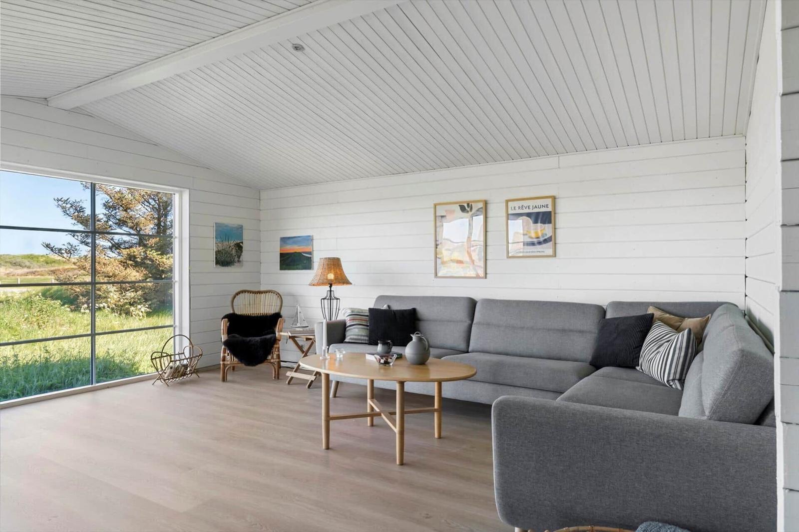 Wohnzimmer mit grauem Sofa, Holztisch und großem Fenster zum Grasland.