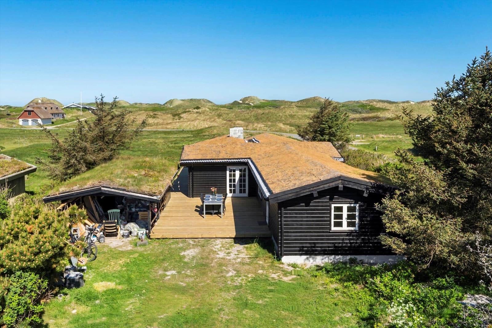 Schwarze Holzhütte mit Grasdach und Terrasse im grünen Landschaftshintergrund.