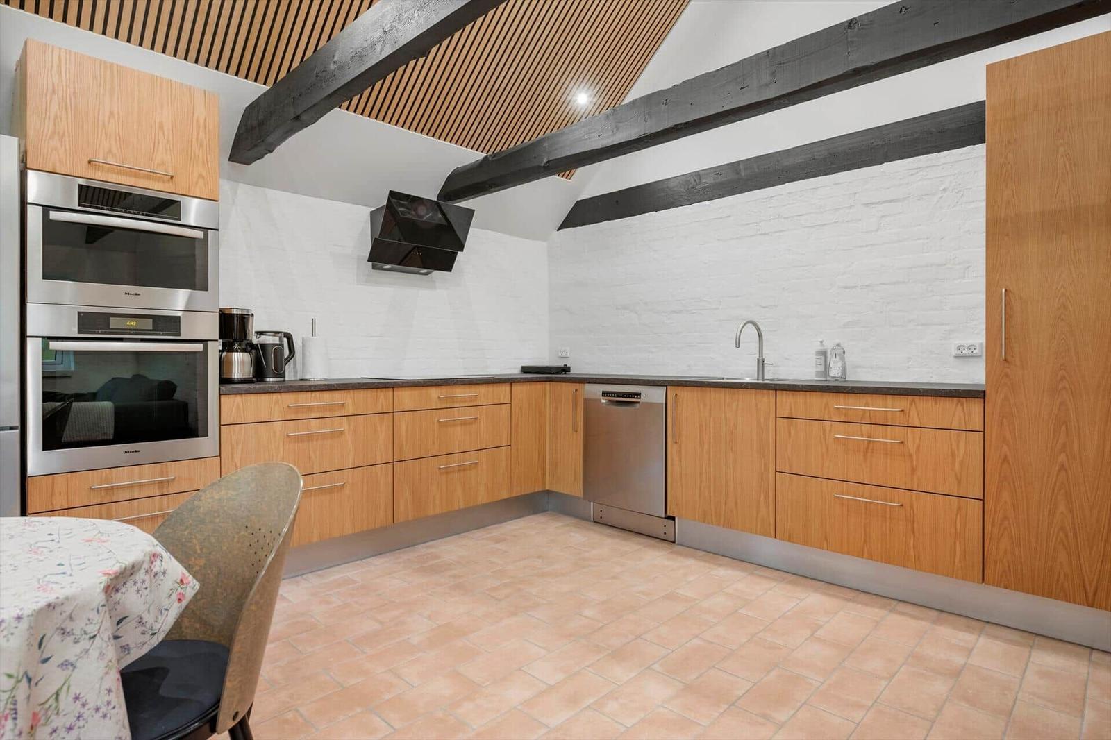 Kitchen with wooden cabinets, stainless steel appliances, and stone floor under sloped ceiling with wooden beams.