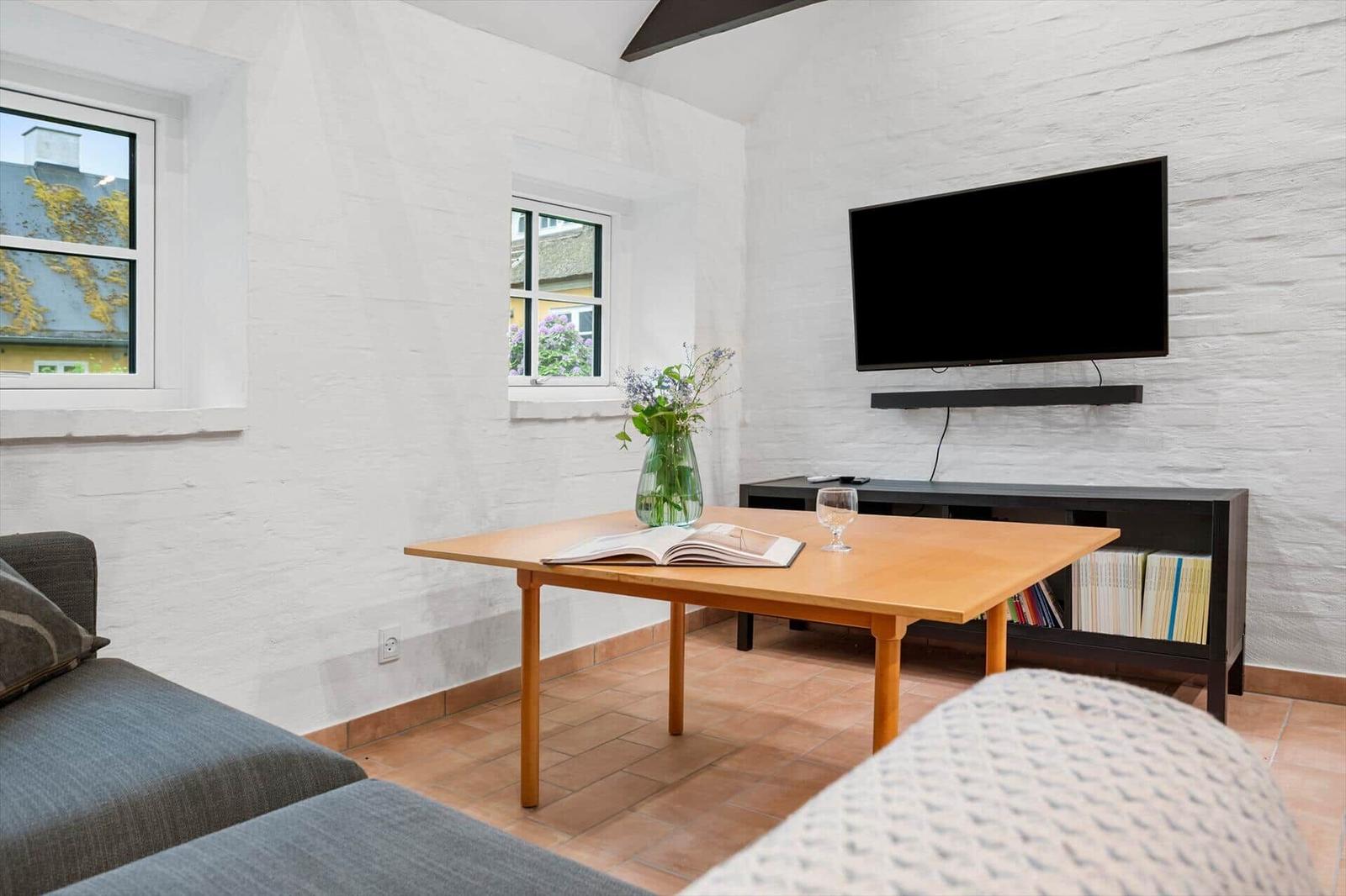 Living room with wooden table, TV, and windows. Bookshelf and flowers on the table.