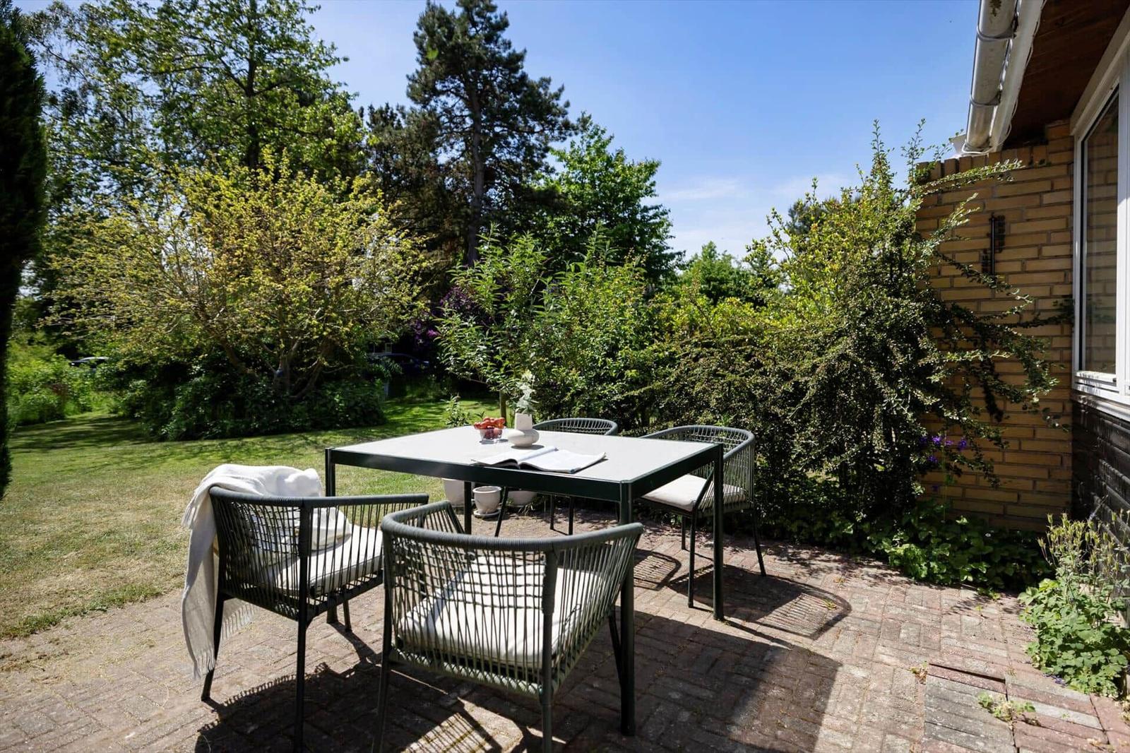 Backyard with table and four chairs, surrounded by greenery and stone paving.