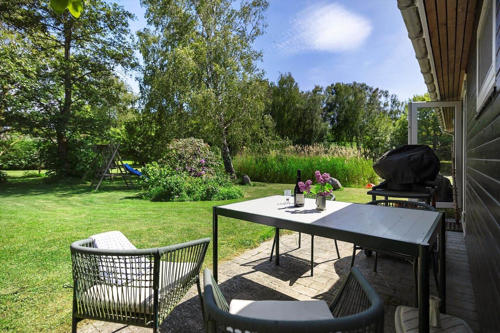 Terrace with table, chairs, and grill. Lawn, trees, and flowers in the background.