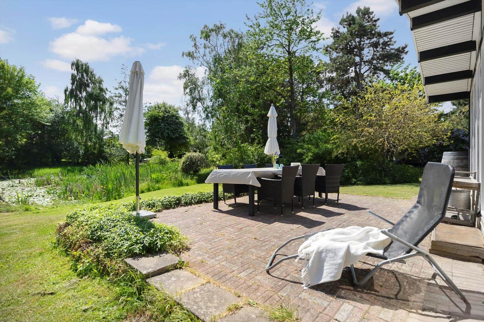 Terrace with table, chairs, and lounge chair. Background: green areas and water.