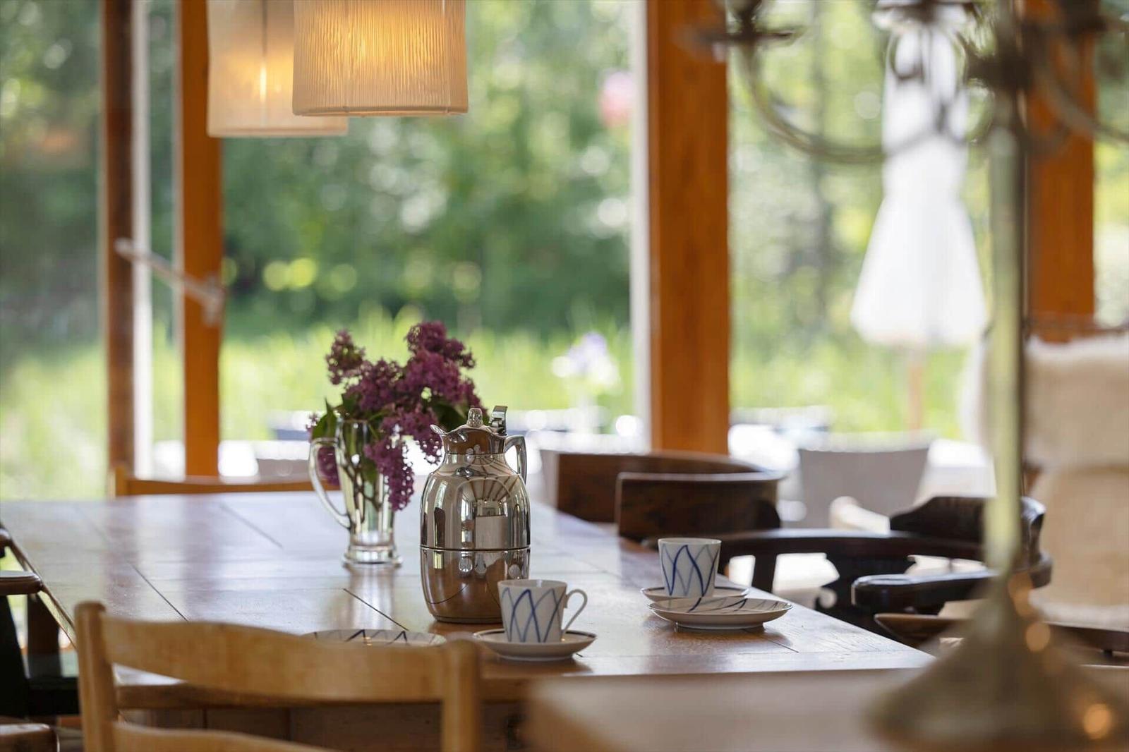 Wooden table with cups, kettle, and flowers by large window.