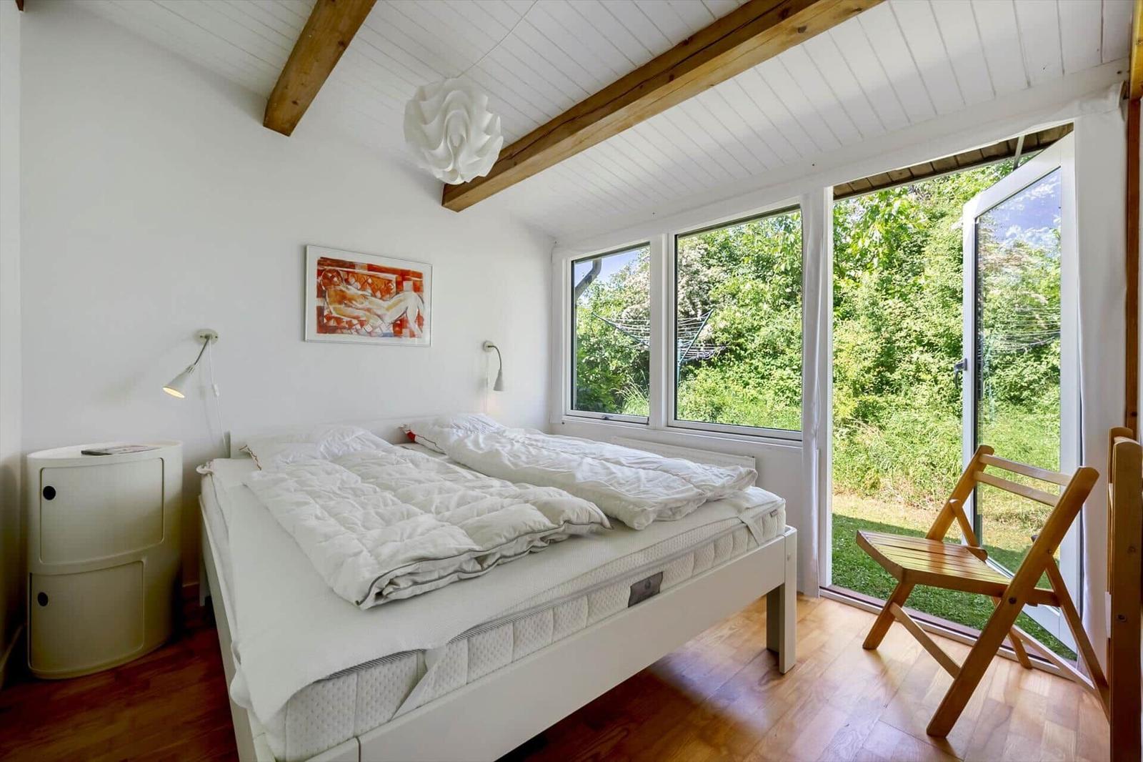 Bedroom with white bed, wooden beams, and view of green outdoor area.