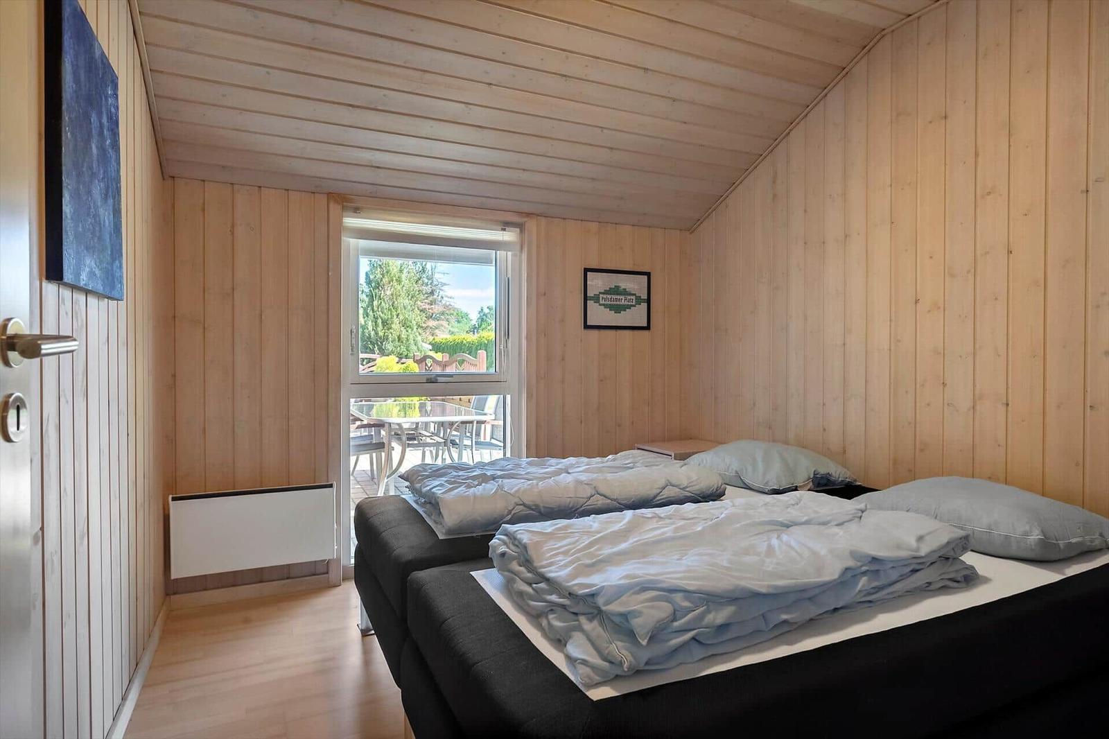 Schlafzimmer mit Holzwand und Bett. Durchgang zur Terrasse mit Tisch und Stühlen.