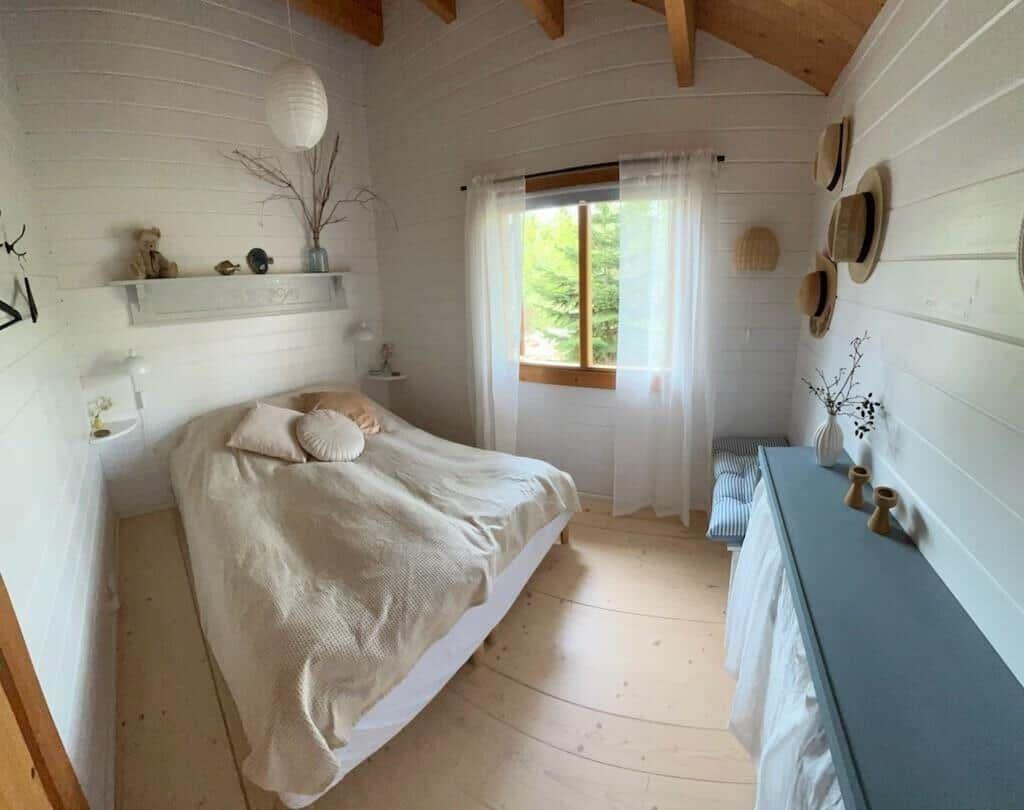 Schlafzimmer mit Bett, Fenster und Holzboden. An der Wand hängen Hüte.