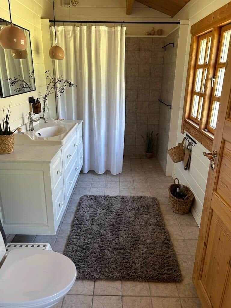 Badezimmer mit Dusche, Waschbecken und Toilette. Holzfenster und Fliesenboden.