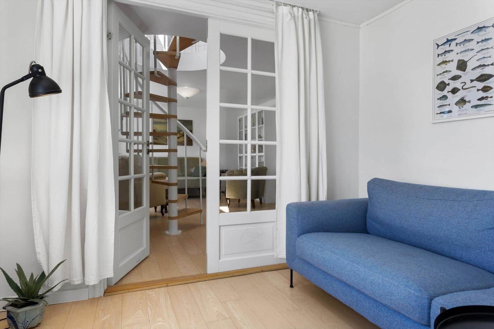 Ein Raum mit blauem Sofa, weißen Vorhängen und einer Tür zum Treppenhaus.