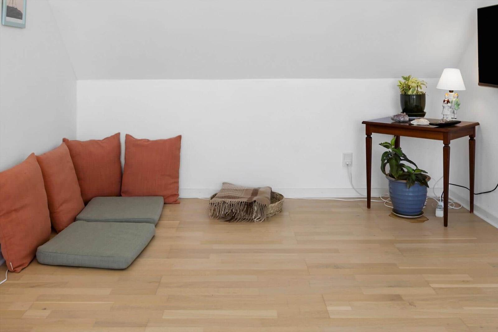 Raum mit Holzboden, orangefarbenen Kissen und grauen Sitzkissen. Tisch mit Pflanzen und Lampe.