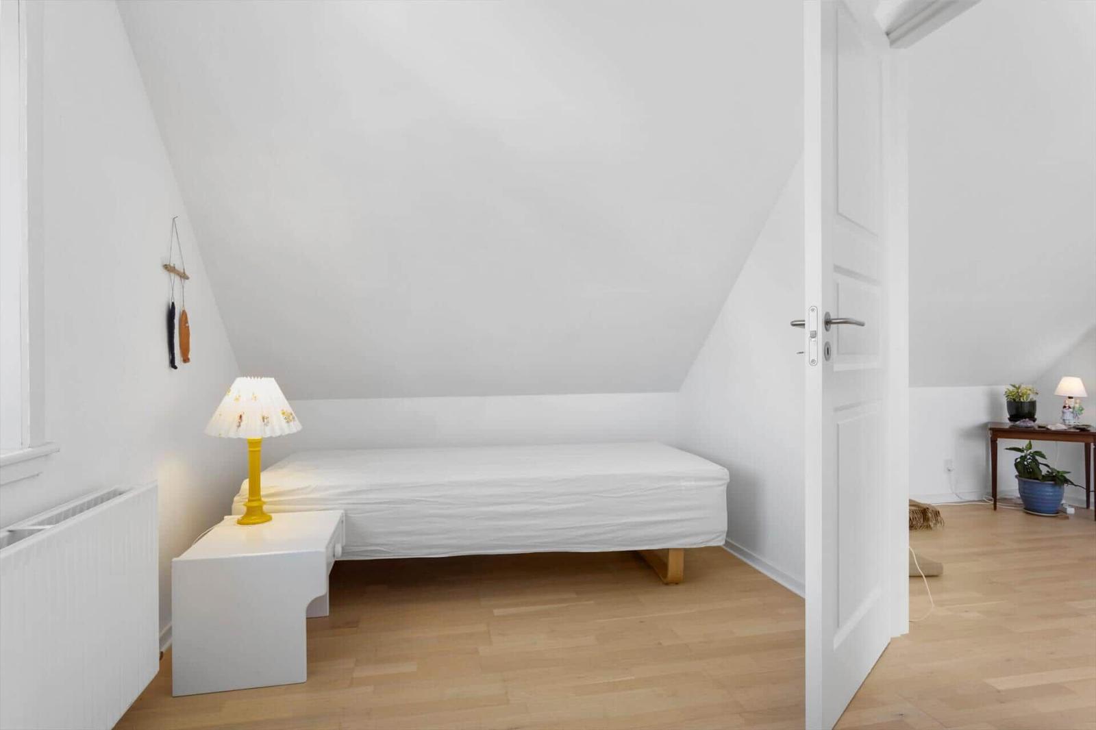 Schlafzimmer mit Dachschrägen, weißem Bett und Nachttisch mit gelber Lampe.