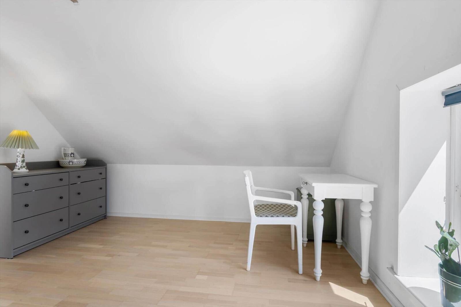 Ein Zimmer mit schrägen Wänden, einem weißen Schreibtisch und Stuhl sowie einem grauen Schrank.