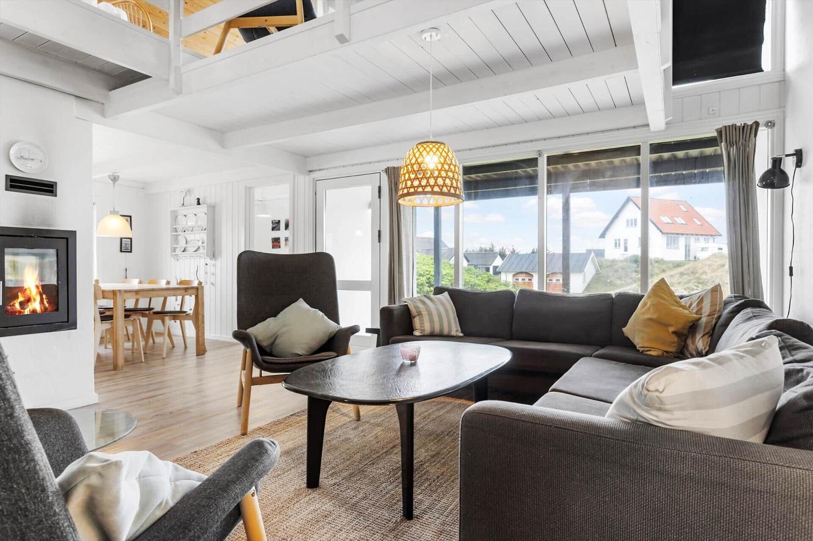 Wohnzimmer mit Sofa, Sessel und Kamin. Durchgehende Fensterfront mit Blick auf Außenbereich.