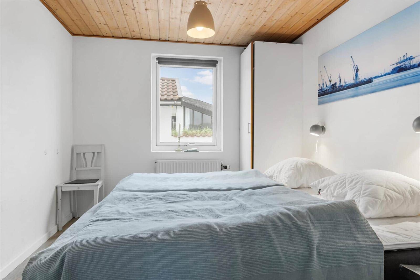 Schlafzimmer mit Bett, Fenster und Holzdecke. Wandbild und Schreibtisch sind sichtbar.