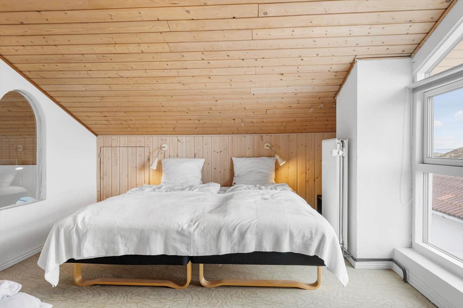 Schlafzimmer mit Doppelbett, Holzdecke und Fenster. Wandlampe und Heizung sichtbar.