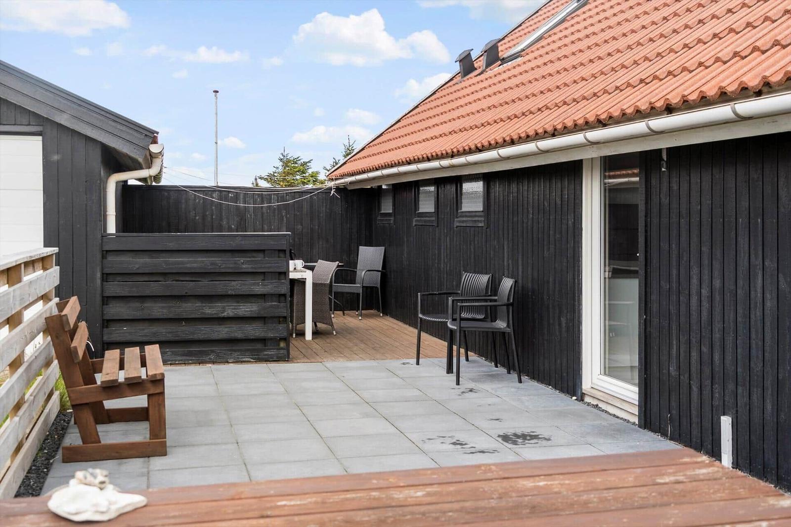 Terrasse mit Tisch, Stühlen und Holzbank vor schwarzem Haus mit rotem Dach.