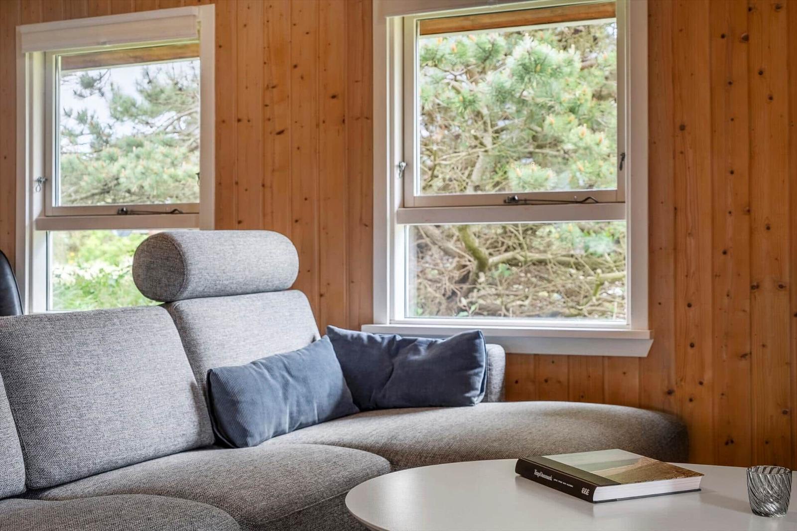 Graue Couch mit Kissen vor Holzwänden und Fenstern mit Blick auf Bäume.