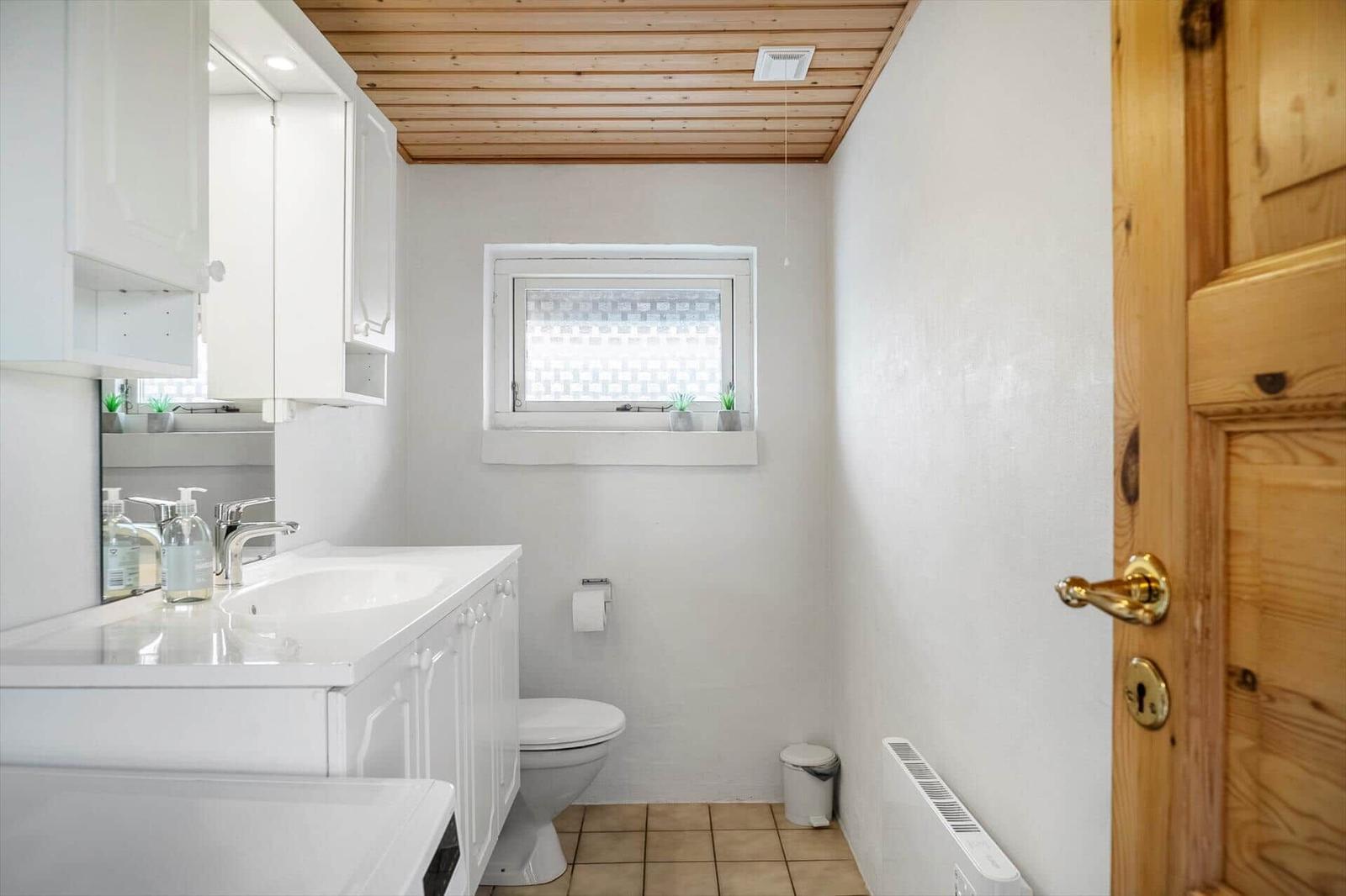 Badezimmer mit Waschbecken, Toilette und Fenster. Holzdecke und Tür.