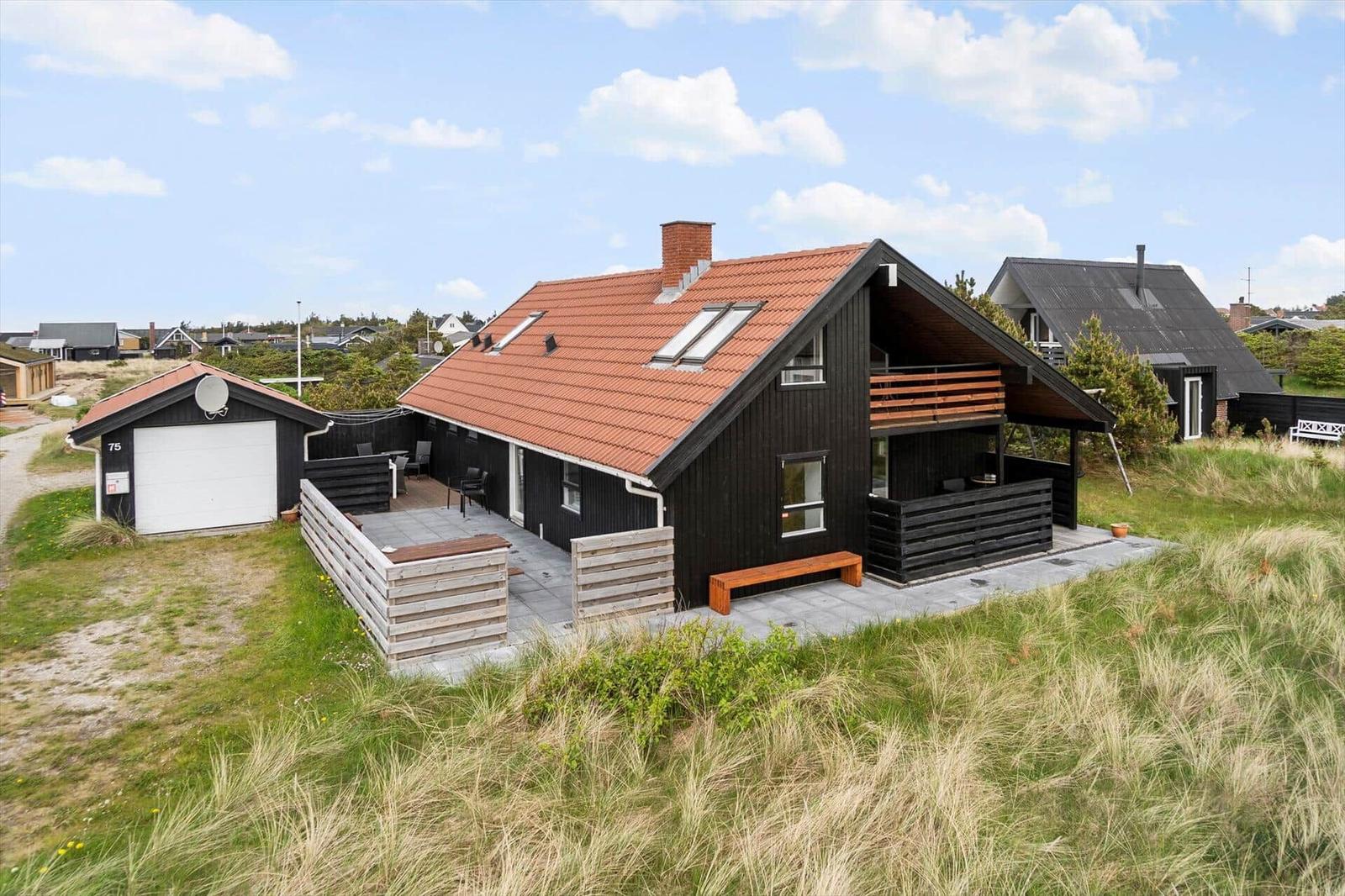 Modernes Haus mit Terrasse, Holzveranda und rotem Dach. Umgeben von Gras und weiteren Häusern.