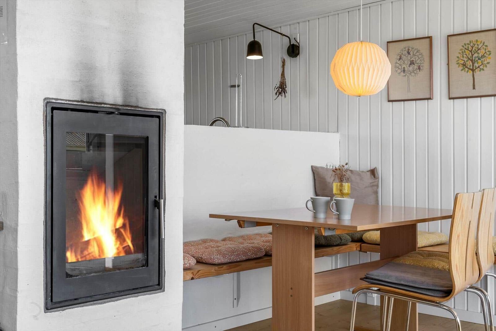 Kamin mit Flammen, Esstisch mit Holzbank und Stühlen, Wand mit Bildern und Pendelleuchte.