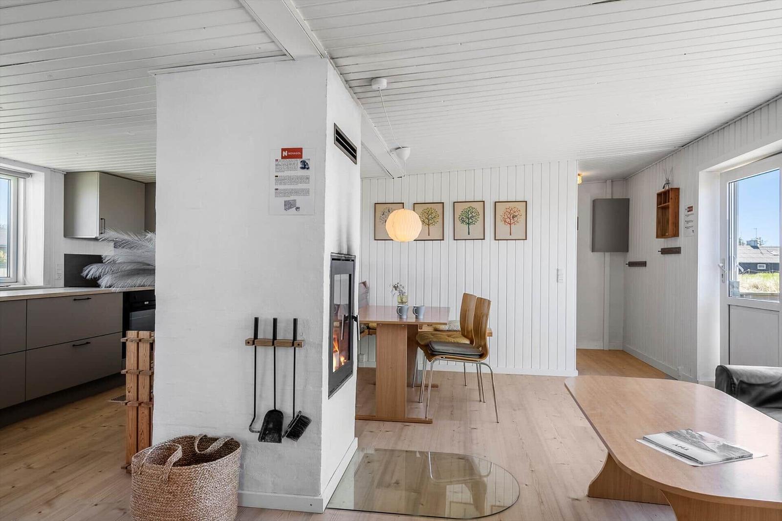 Küche, Essbereich und Wohnbereich mit Holzboden und weißer Wandverkleidung. Kamin und Tisch mit Stühlen sichtbar.