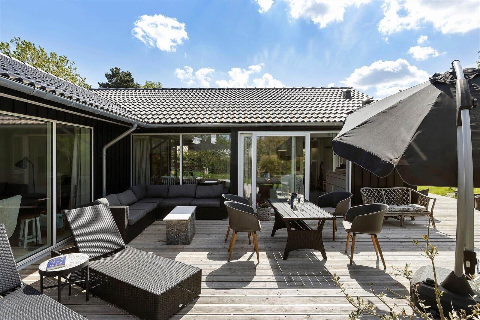 Deck with seating group, table, chairs, and umbrella. House with glass facade and roof.