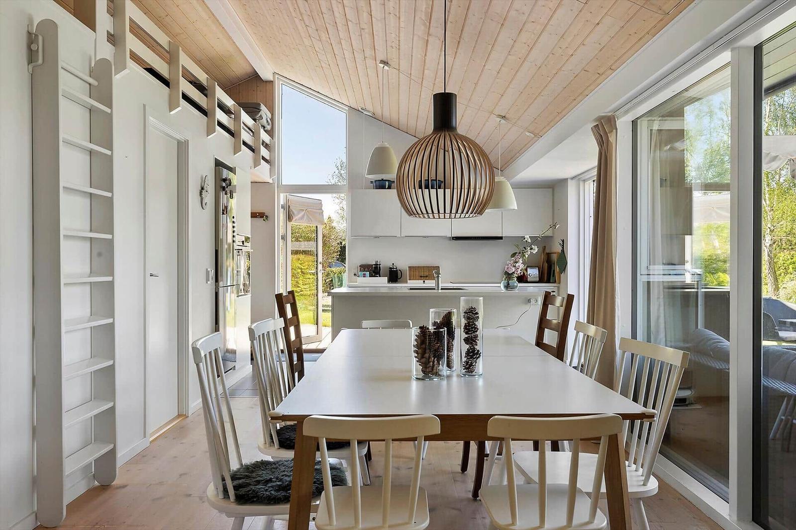 Dining area with table, chairs, and kitchen. Wooden ceiling and large windows.