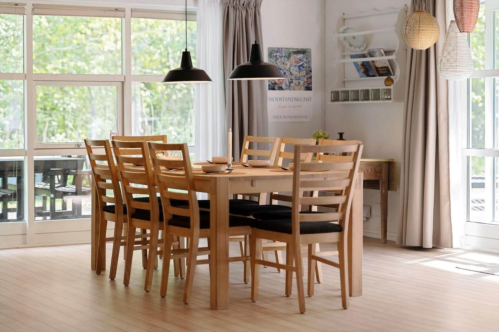 Dining room with wooden table, chairs, and large windows. Hanging lamps and wall shelf are visible.