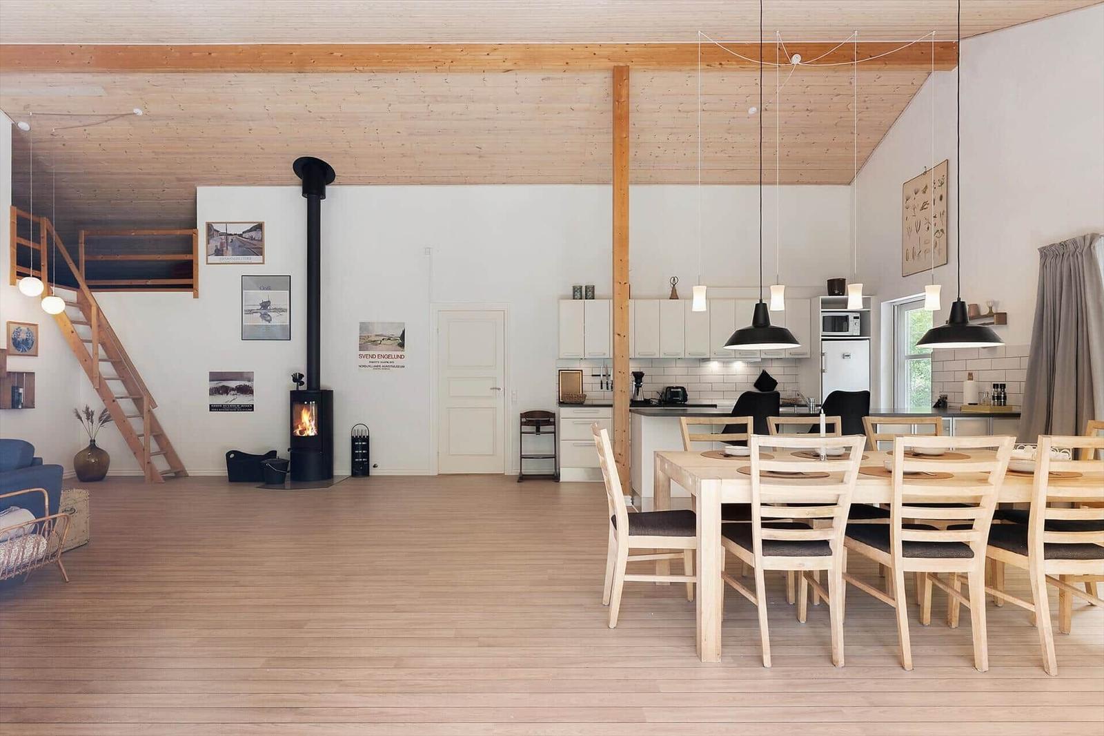A room with dining area, kitchen, and fireplace. Wooden floor and ceiling beams.