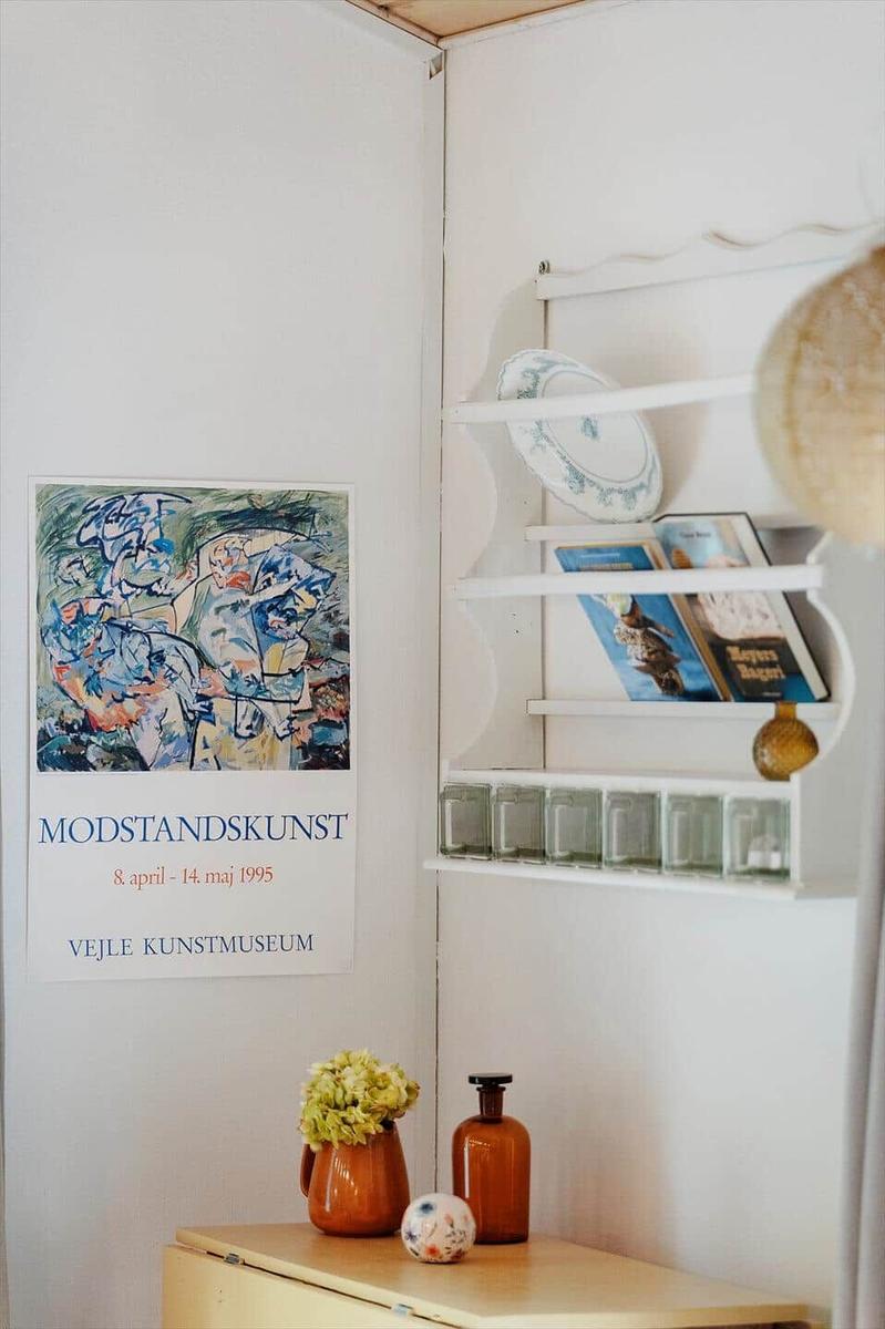Wall with art poster, shelf with plates and books, wooden surface with vase and bottle.