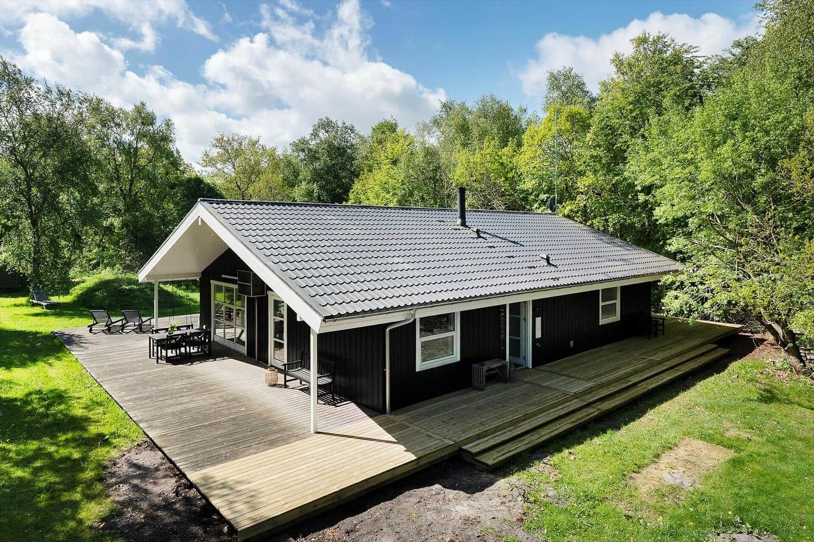 Wooden house with terrace and garden, surrounded by trees under blue sky.