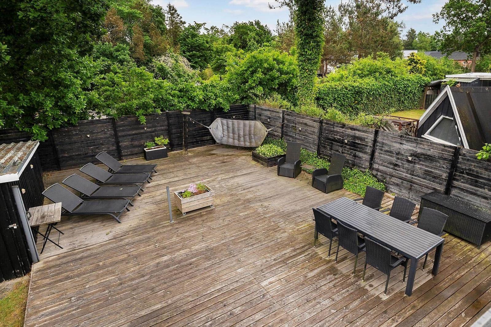 Holzterrasse mit Liegestühlen, Esstisch und Hängemattenschaukel umgeben von grüner Hecke.