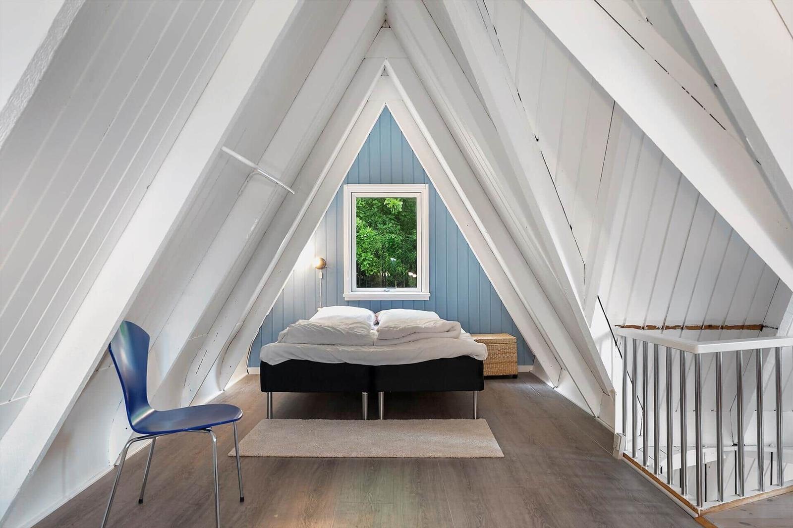 Schlafzimmer mit Dachschrägen, blauer Wand und Fenster. Bett, Stuhl und Teppich sind sichtbar.