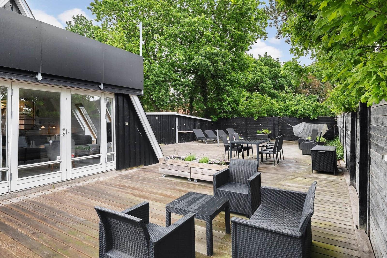 Terrasse mit Gartenmöbeln, Tisch, Liegen und Hängematten. Holzdeck und grüne Bäume im Hintergrund.