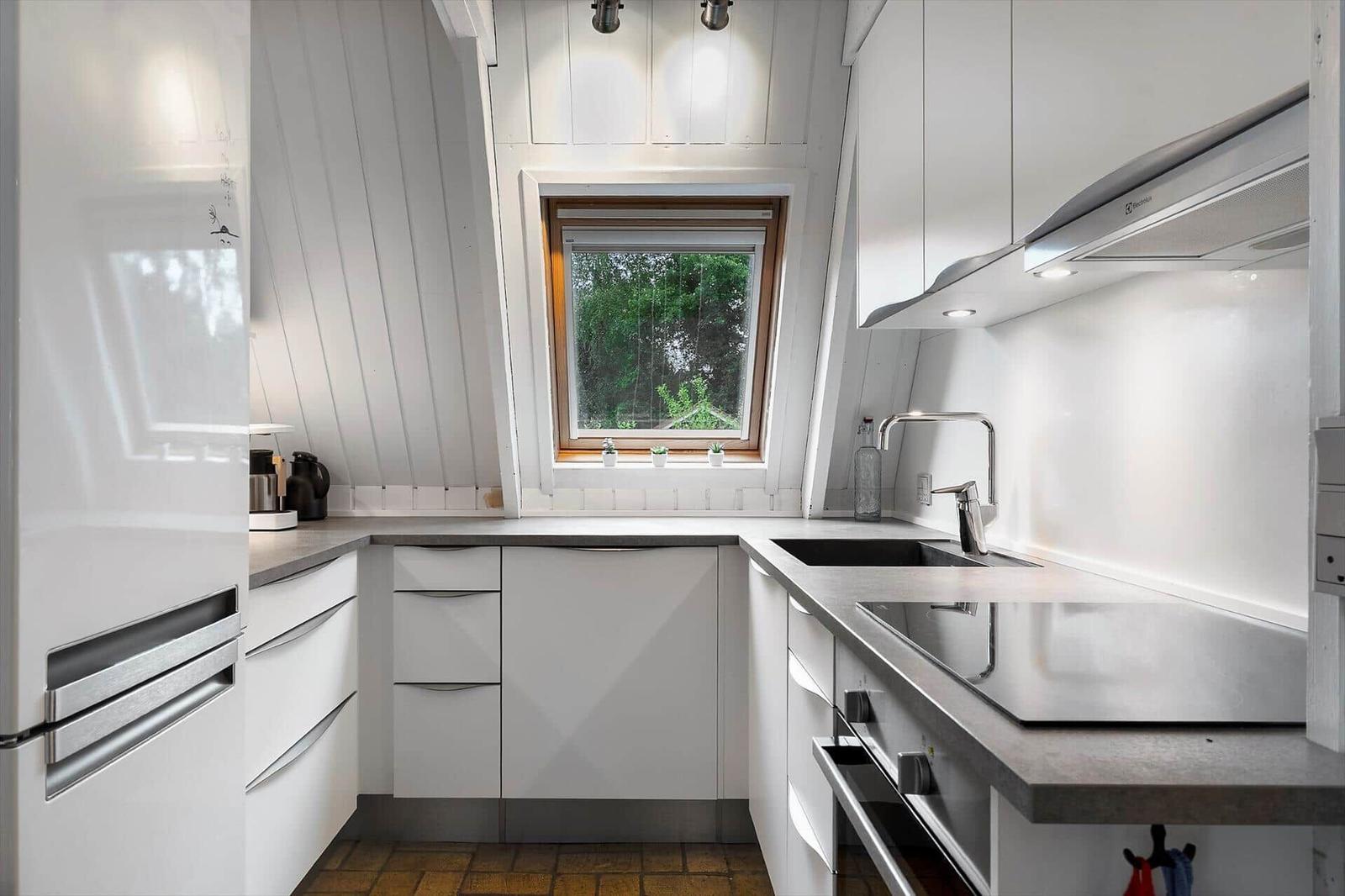 Küche mit weißen Schränken, Arbeitsplatte und Fenster. Kühlschrank und Spüle sichtbar.