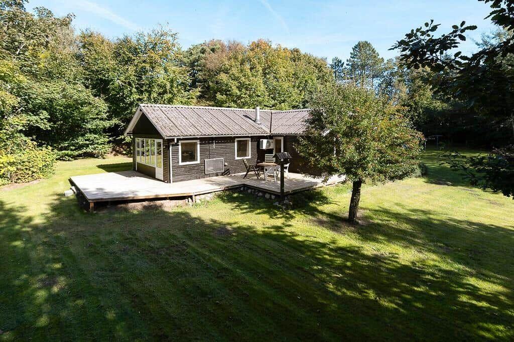 Holzhütte mit Terrasse und Garten. Umgeben von Bäumen unter blauem Himmel.