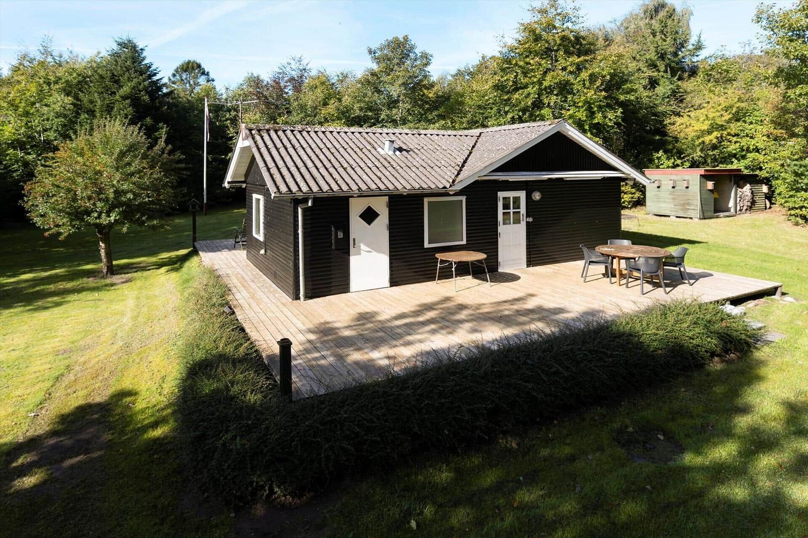 Schwarzes Holzhaus mit Terrasse, Tisch und Stühlen. Umgeben von Gras und Bäumen.