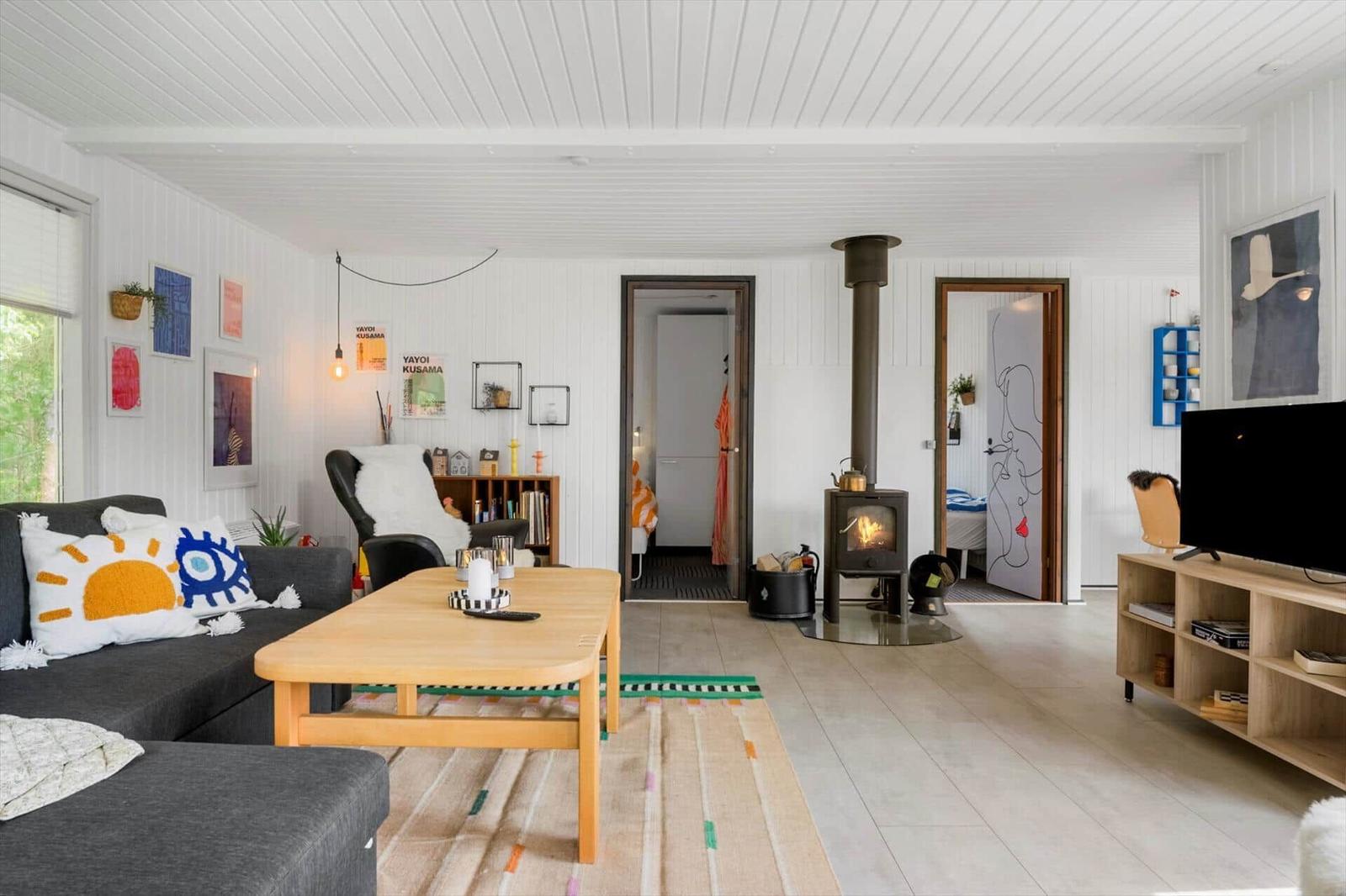Wohnzimmer mit Kamin, Sofa und Fernseher. Holztisch, Wandbilder und Fenster sichtbar.