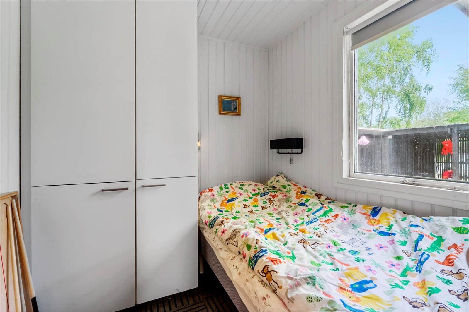 Ein Schlafzimmer mit Bett, Kleiderschrank und Fenster. Auf dem Bett liegt ein buntes Bettlaken.