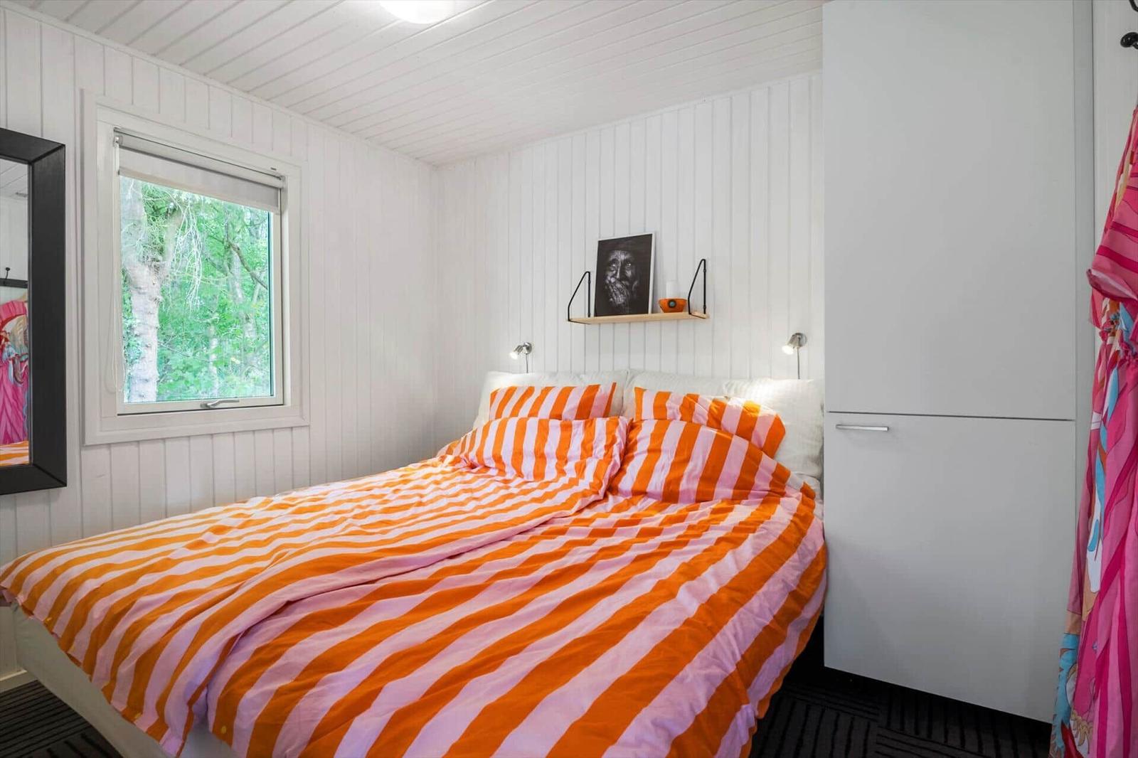Schlafzimmer mit Bett, Fenster und Schrank. Bett mit orangefarbenem Bettzeug.