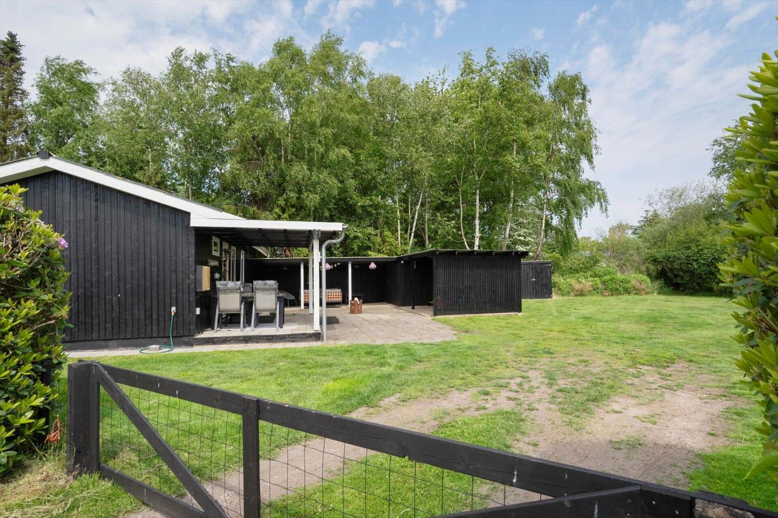 Schwarzes Holzhaus mit Terrasse, Garten und Bäumen im Hintergrund.