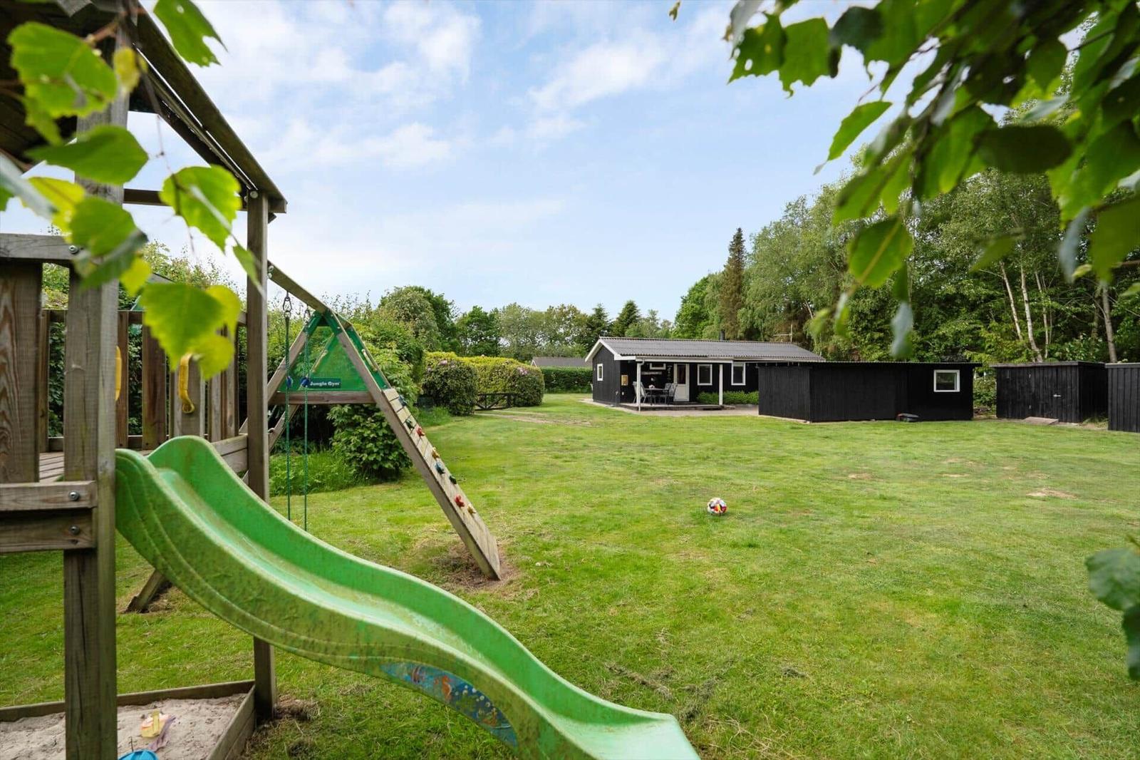 Grüner Rutschbahn im Garten, Ferienhaus mit Terrasse und umliegenden Bäumen.