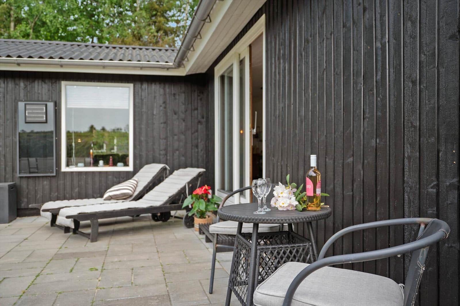 Terrasse mit Liegen, Tisch und Stühlen. Flasche Wein und Blumen darauf. Dunkler Holzaufbau.