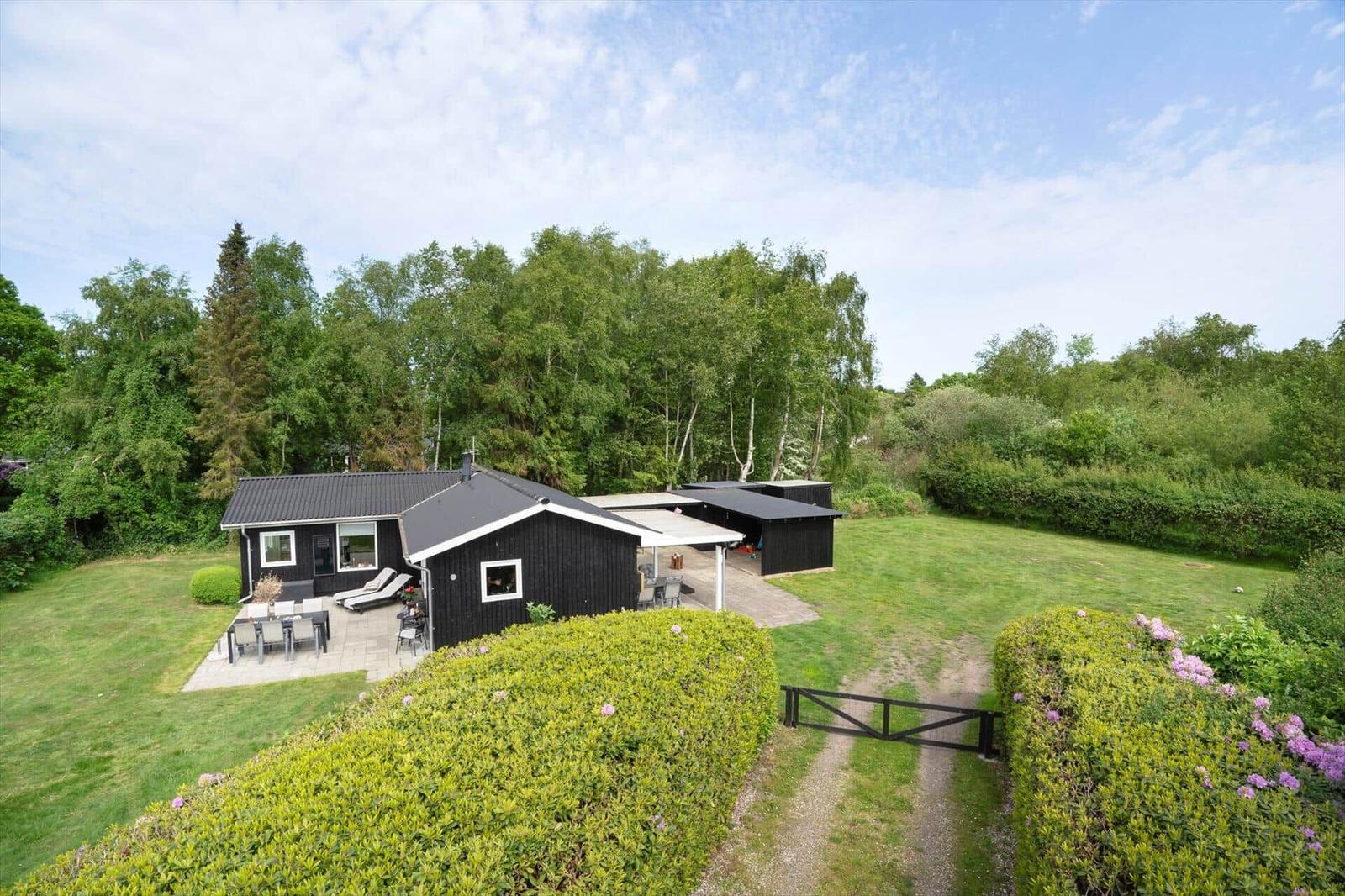 Schwarze Holzhütte mit Garten, Terrasse und Grillplatz. Umgeben von Bäumen und grünem Rasen.