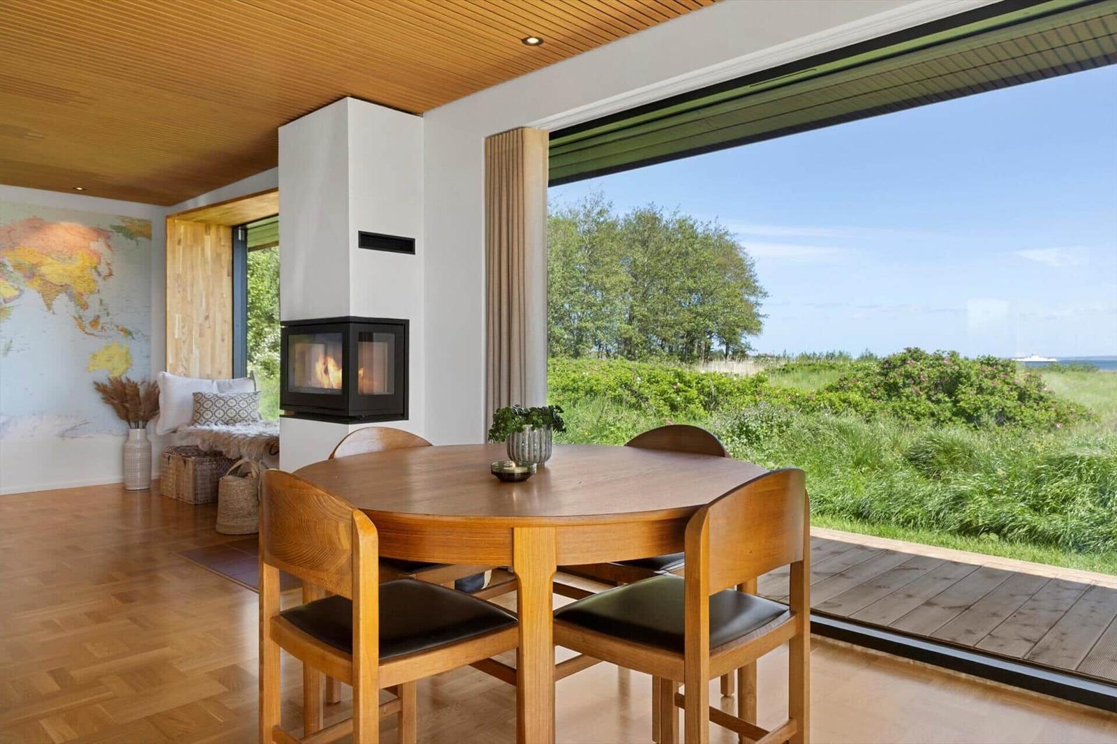 Dining area with wooden table, seating, and fireplace. Large windows overlooking green land and sea.
