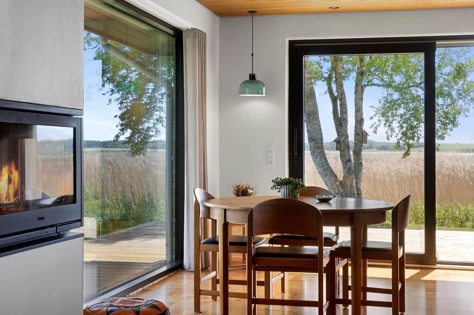 Dining area with table and chairs, large windows overlooking forest and lake, fireplace in background.