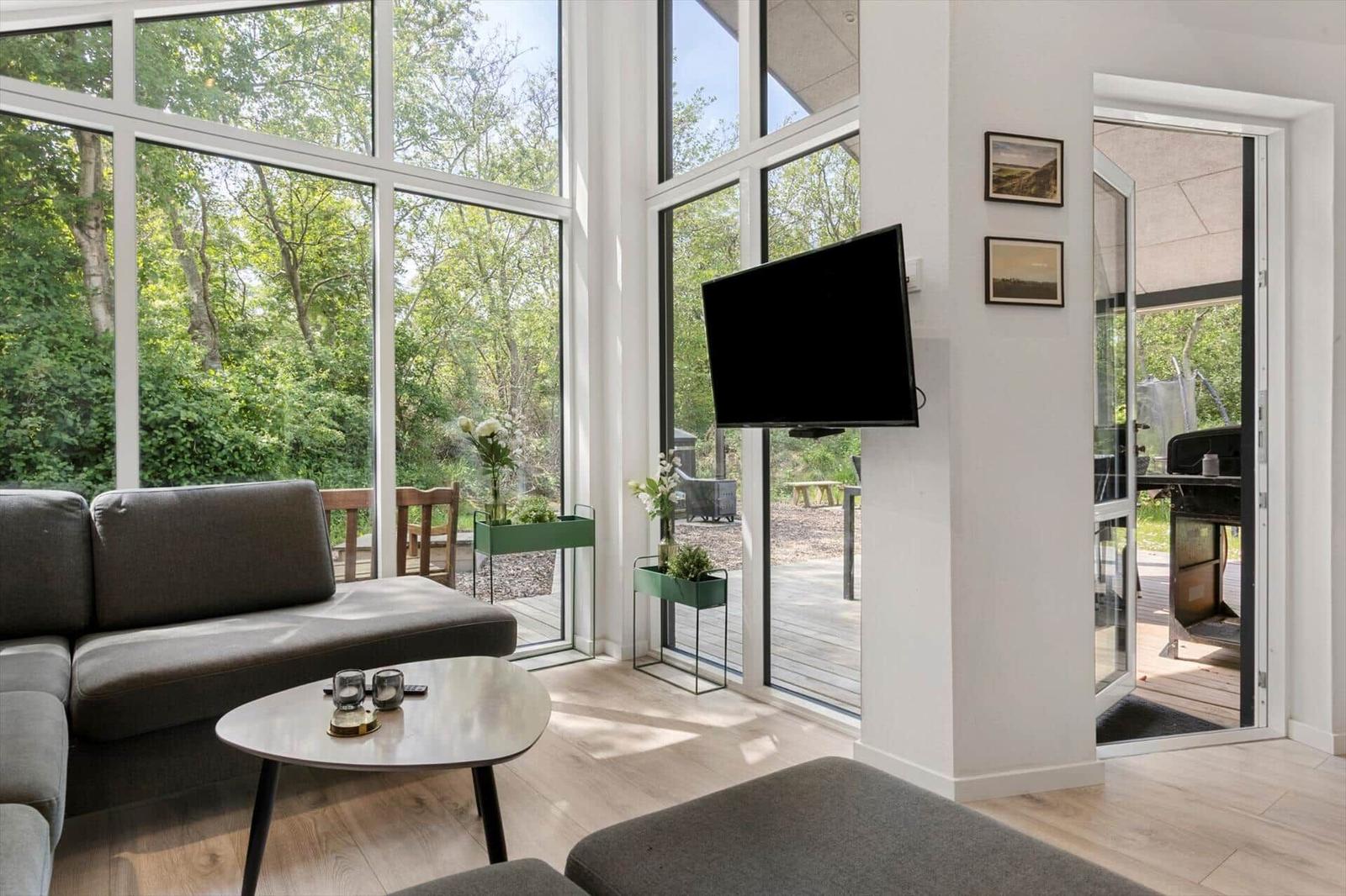 Wohnzimmer mit großem Fenster, Sofa, Couchtisch und Fernseher. Durchgang zur Terrasse mit Grill.